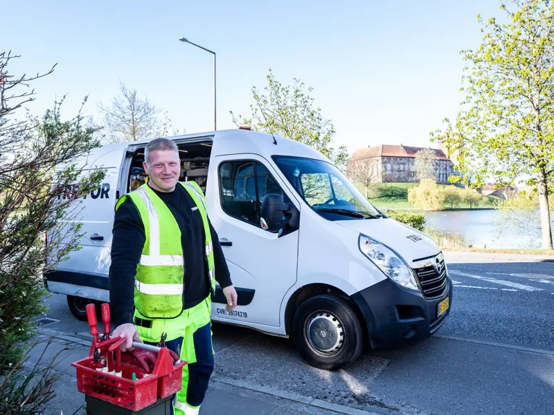 Smiling worker in reflective vest stands by a van holding tools.