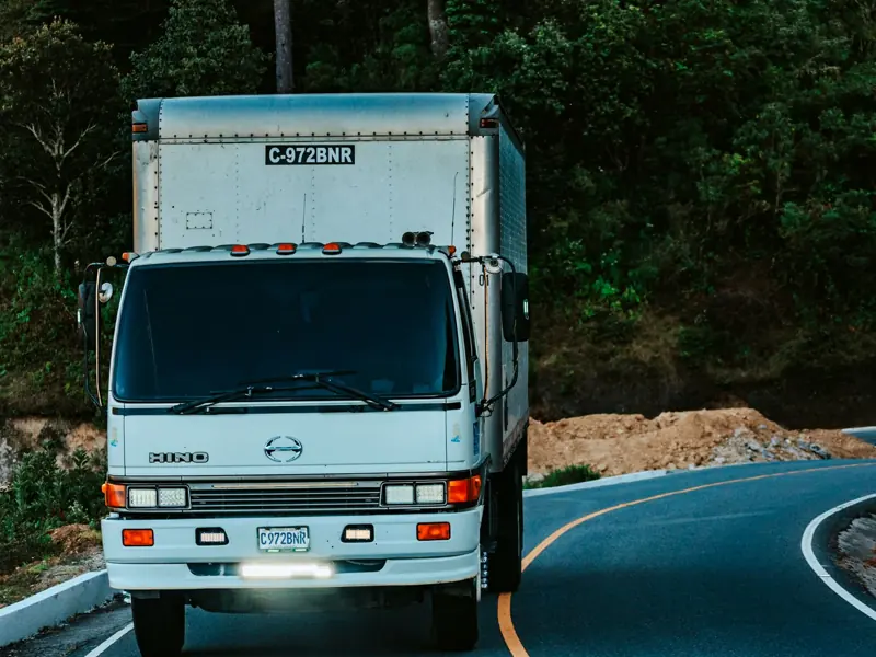 A white truck driving on a winding road surrounded by dense green forest plants.