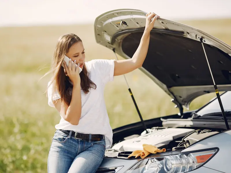 Worried young female driver in white t shirt and jeans speaking on phone while leaning on broken car with open hood asking for help with repair against green field in sunny summer day