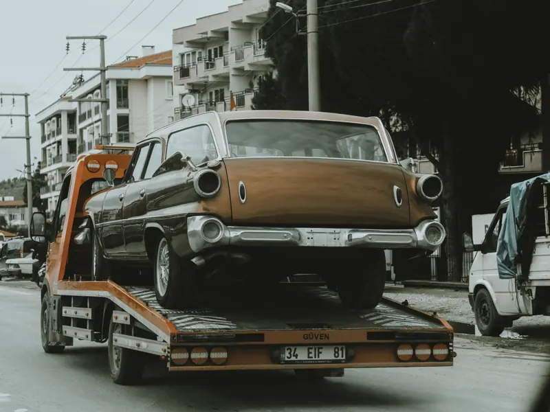 A vintage car loaded on a tow truck in a city street, surrounded by modern buildings.