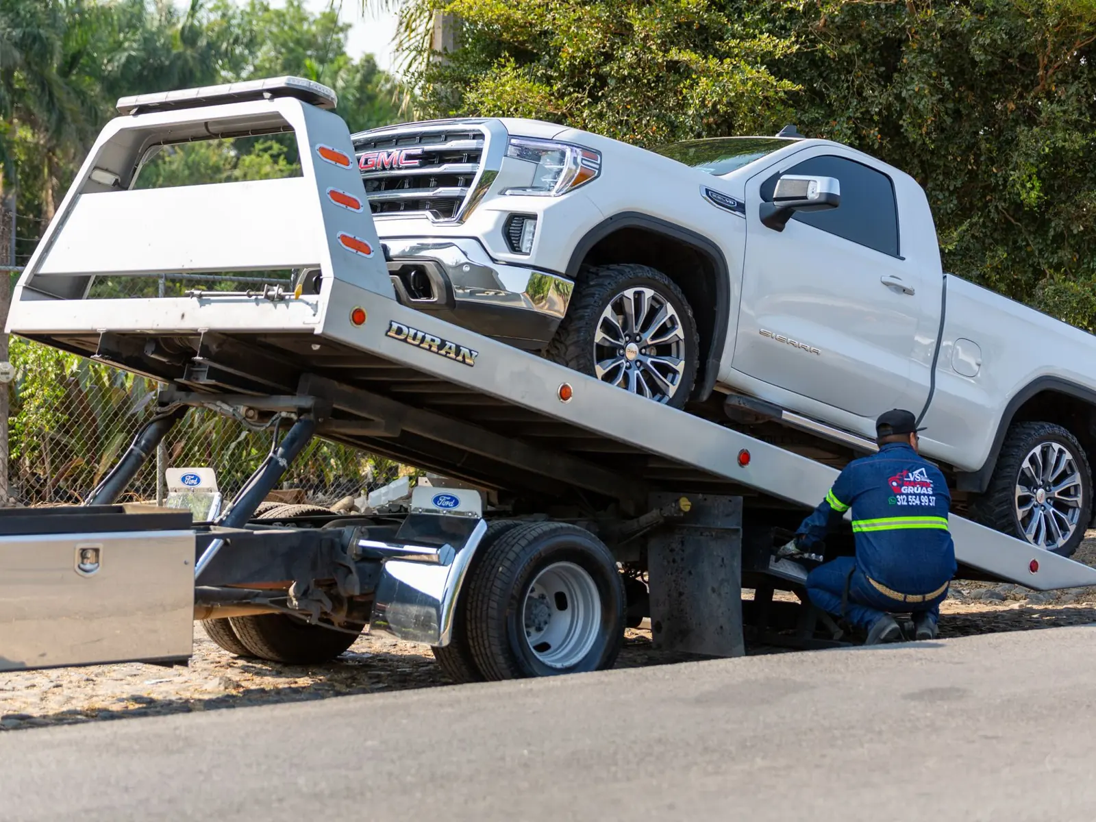Tow truck operator loading white GMC pickup truck on street in daytime.