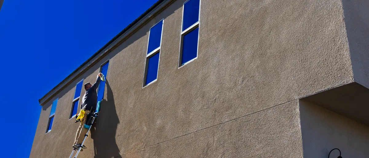 Man using a ladder to clean exterior windows of a tall building under a clear blue sky.
