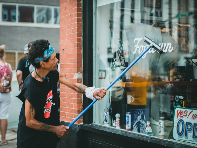 A man with tattoos cleaning a glass storefront window on a busy street using a squeegee.
