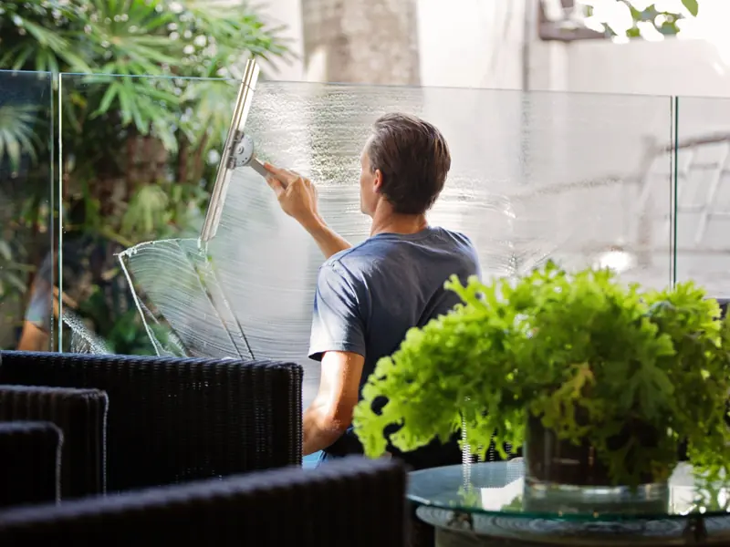 A man cleaning a glass barrier outdoors with a squeegee, surrounded by greenery.