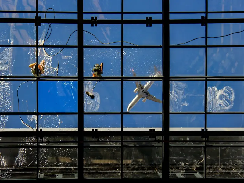 Window cleaners at work with an airplane flying overhead against a blue sky.